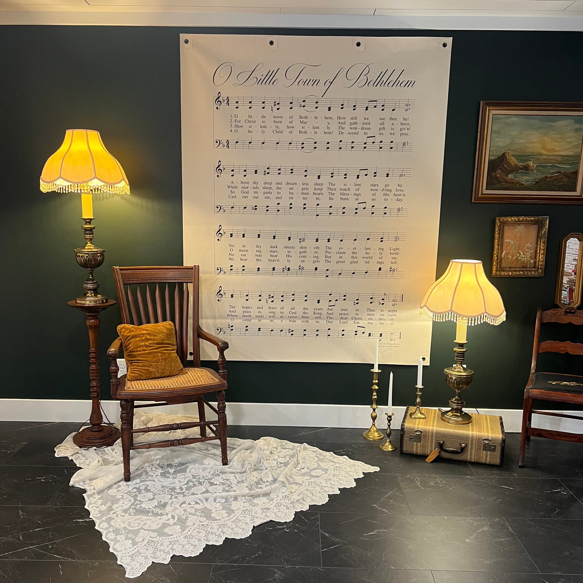 A large vintage-style sheet music wall hanging reading 'O Little Town of Bethlehem' is displayed on a dark green wall in a styled room with a wooden chair, two lit table lamps, a lace rug, and framed wall art.