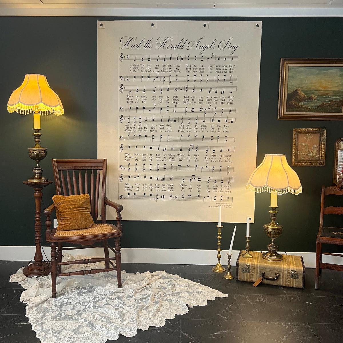 A large wall poster featuring sheet music for 'Hark The Herald Angels Sing' displayed in a styled room with a dark green wall, vintage lamps, a wooden chair, and a lace rug.