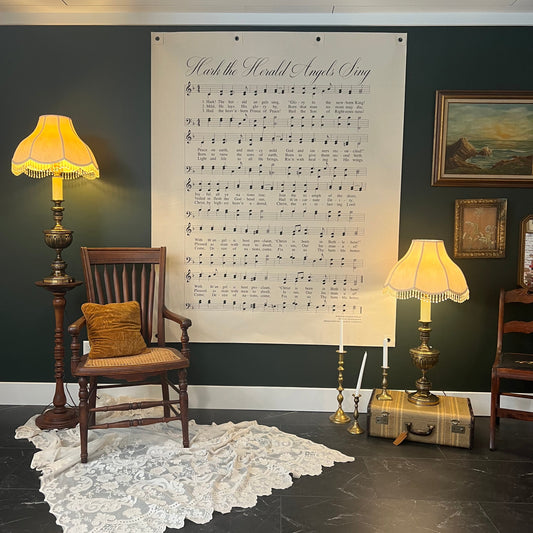 A large wall poster featuring sheet music for 'Hark The Herald Angels Sing' displayed in a styled room with a dark green wall, vintage lamps, a wooden chair, and a lace rug.