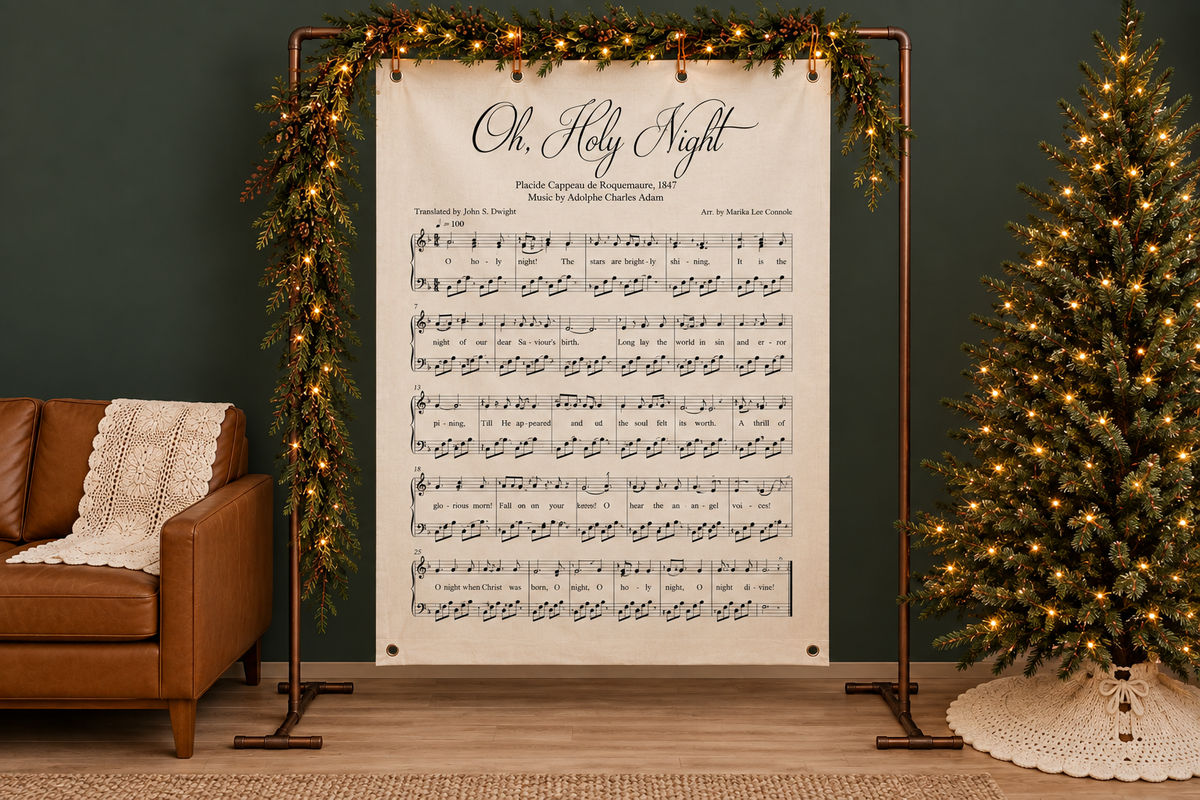 Oh Holy Night Christmas hymn wall canvas with musical notation and lyrics, displayed on a copper frame decorated with green garland and warm white string lights beside a brown leather armchair.
